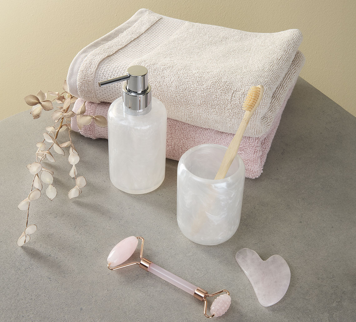 Serviette couleur sable et rose à côté d’un pot à brosse à dents, un distributeur à savon, et rouleau pour le visage